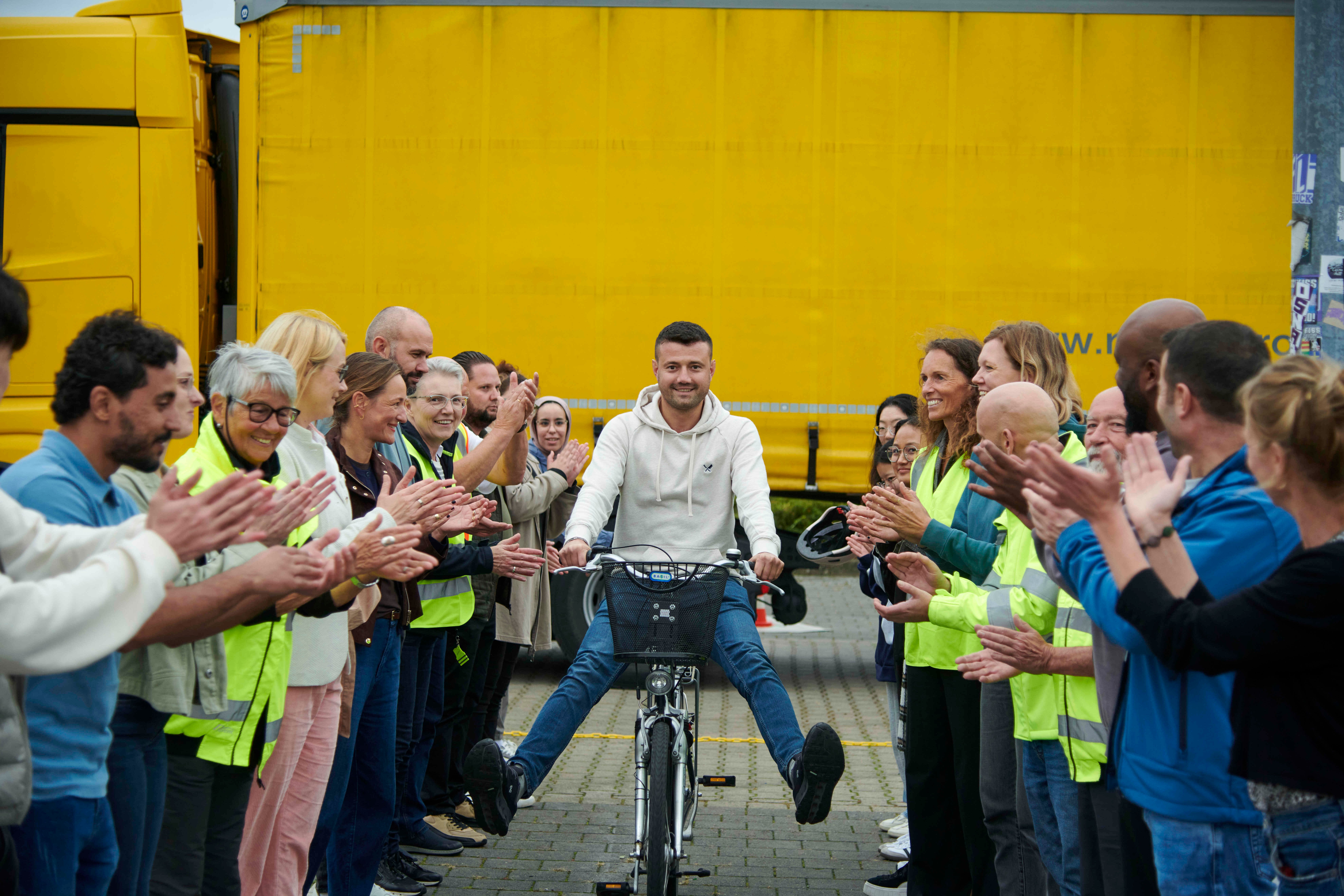 Ein Mann sitzt lächelnd auf einem Fahrrad und wird von einer Gruppe applaudierender Menschen in Warnwesten empfangen. Im Hintergrund steht ein großer gelber Lkw.