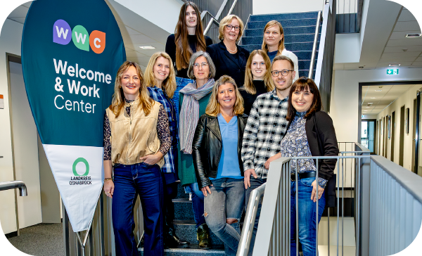 Das Beratungsteam des Welcome and Work Centers (WWC) Landkreis Osnabrück lächelt auf einer Treppe. Links steht ein großer, blauer Aufsteller mit dem WWC-Logo, dem Namen der Institution und dem Logo des Landkreises Osnabrück. Das Beratungsteam des Welcome and Work Centers (WWC) Landkreis Osnabrück lächelt auf einer Treppe. Links steht ein großer, blauer Aufsteller mit dem WWC-Logo, dem Namen der Institution und dem Logo des Landkreises Osnabrück.